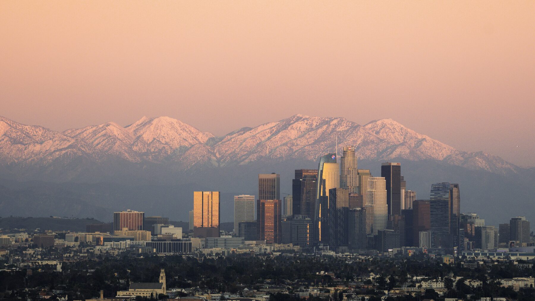 The downtown Los Angeles skyline in front of snow-covered mountains under a pink sky.