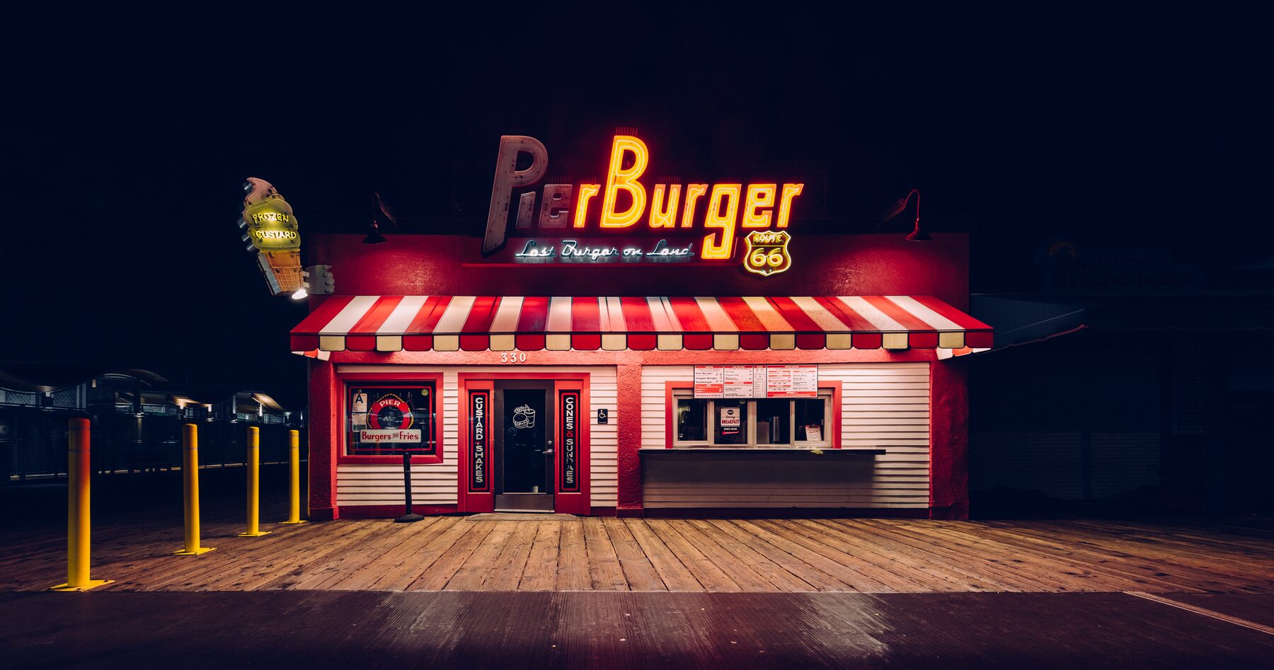 A red and white burger stand glowing under neon on a dark pier.