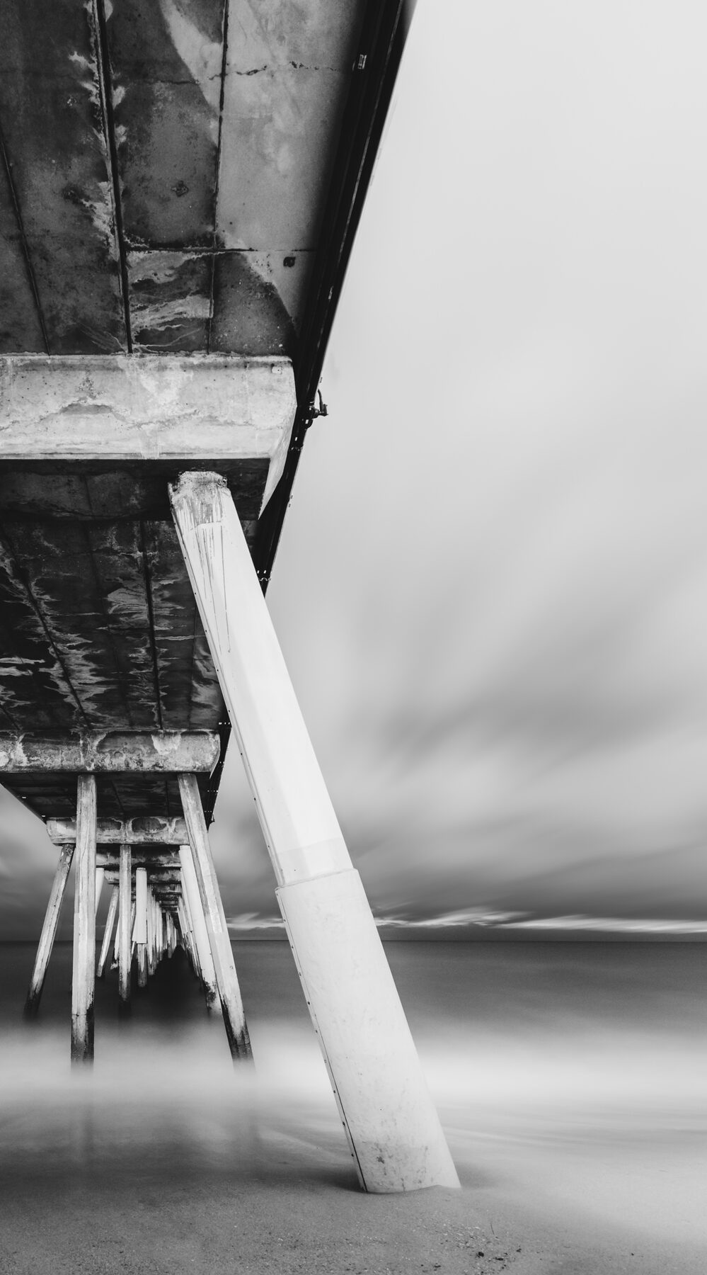 Long-exposure black and white view beneath a pier, the sea smoothed into mist.