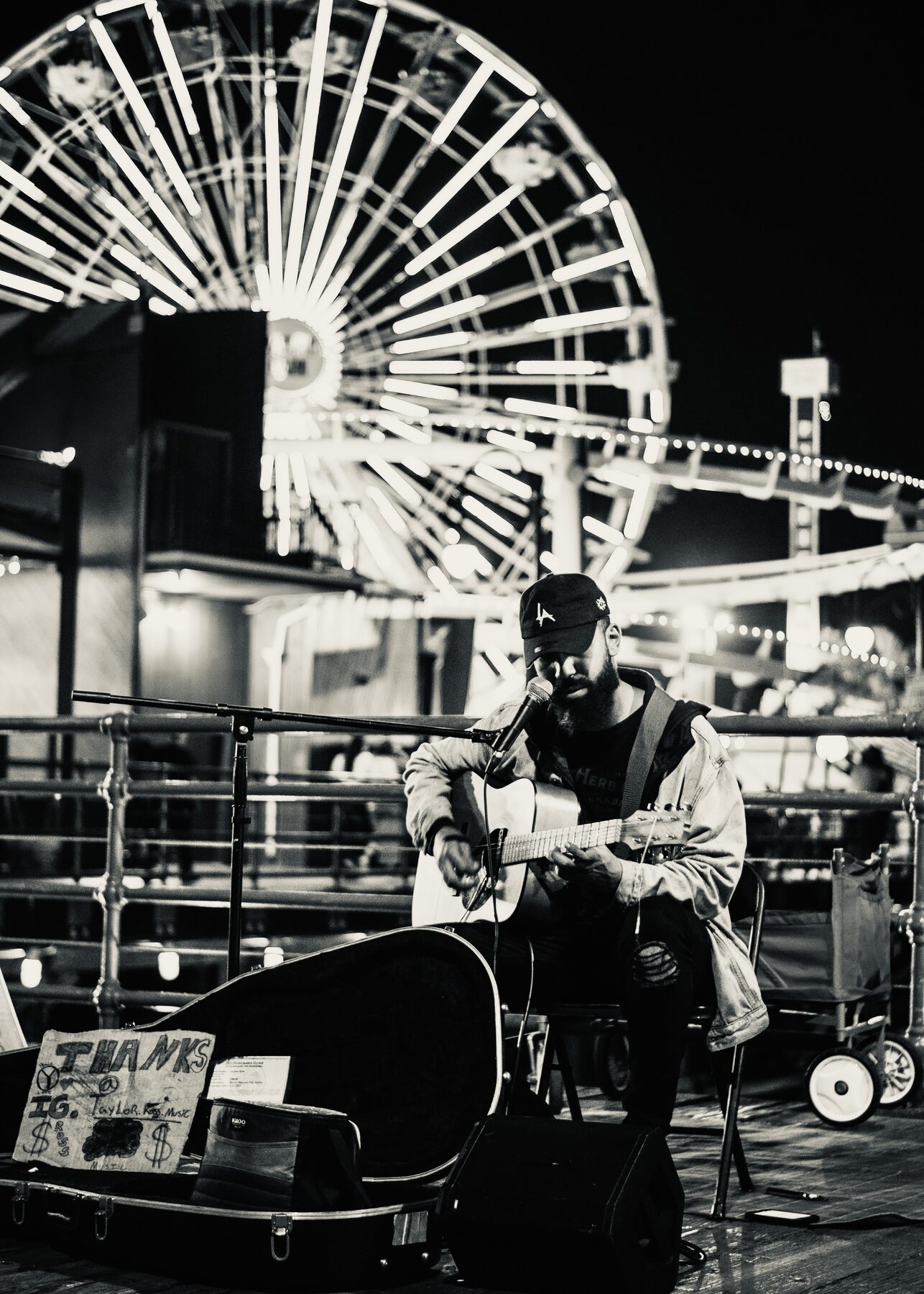 A guitarist playing on a pier at night beneath a lit Ferris wheel, in black and white.