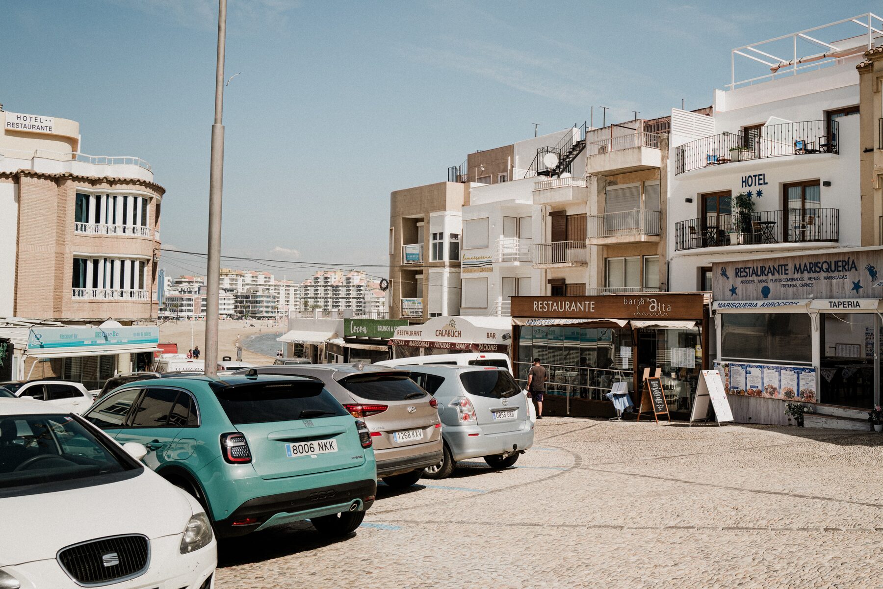 White hotels around a paved square with small cars parked in the sun.