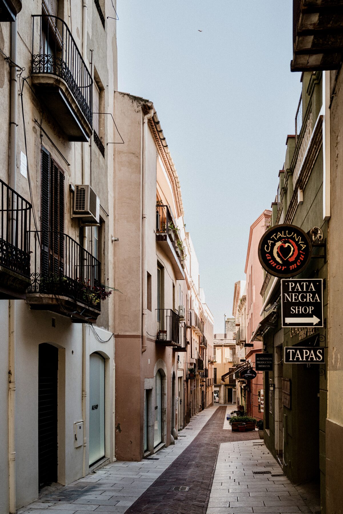 A narrow sunlit alley between tall buildings, a tapas sign hanging on the right.