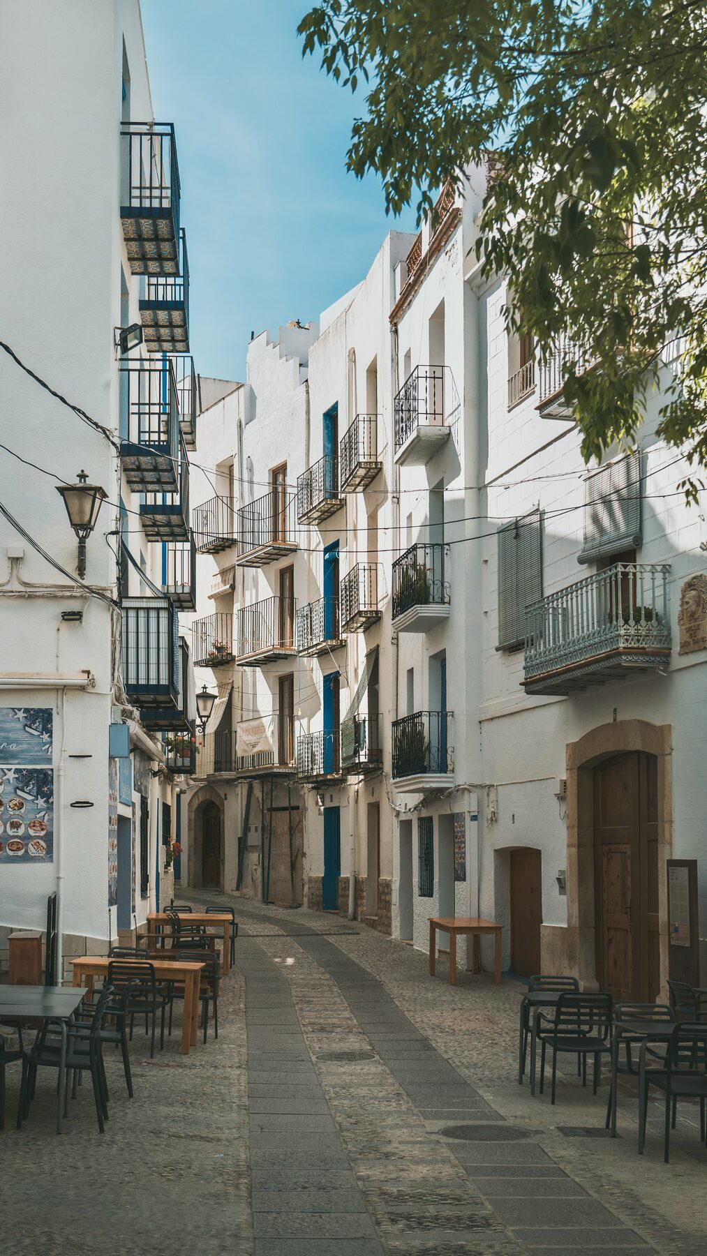 A quiet street of whitewashed houses with café tables and a tree overhead.