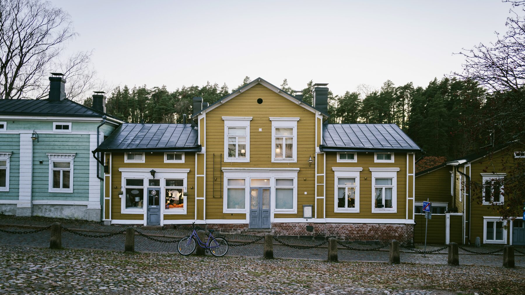 A yellow wooden house with a grey roof beside a pale blue neighbour on a cobbled square.