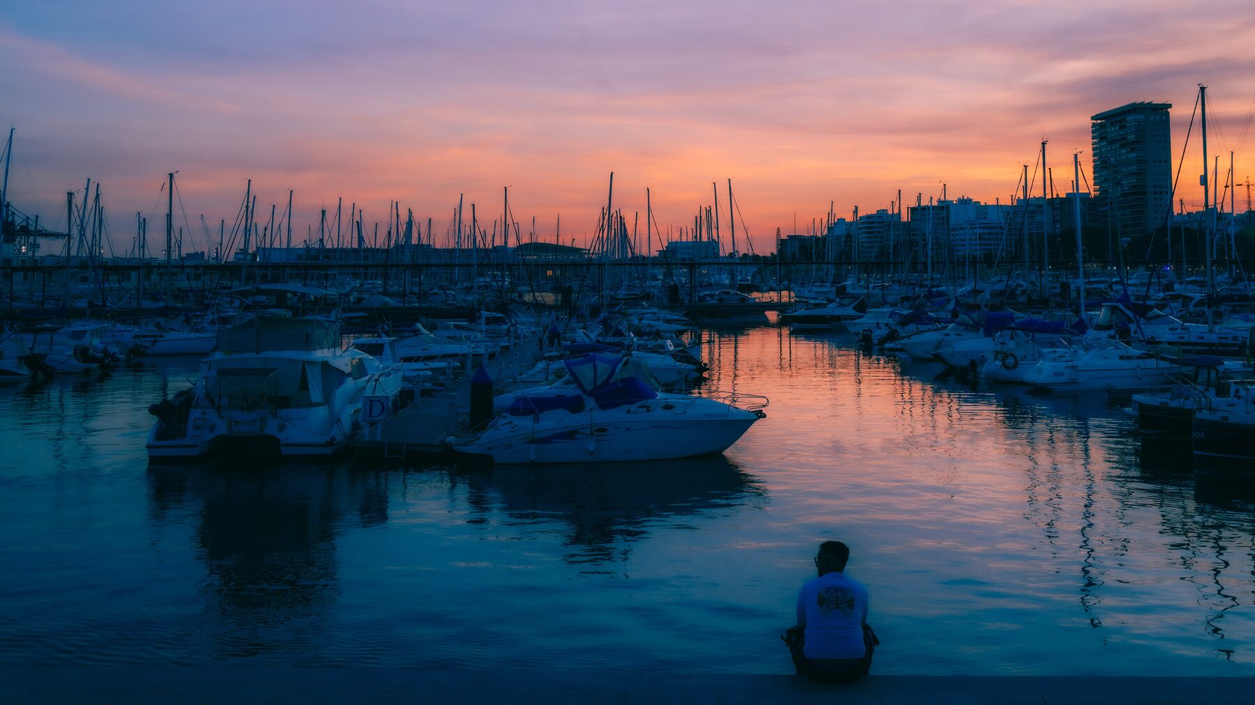 A marina under a pink and violet sunset, one person sitting at the water's edge.