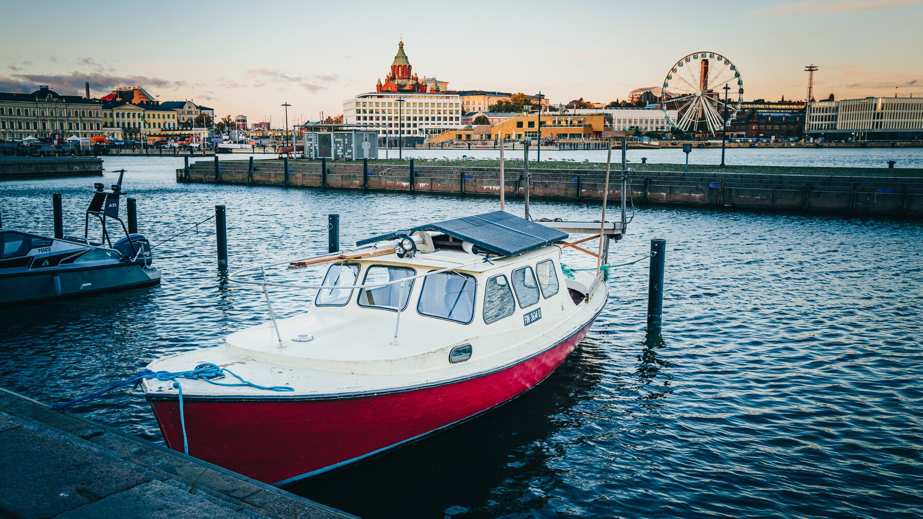 A red and white motorboat moored in a harbour, a Ferris wheel and domed building behind.