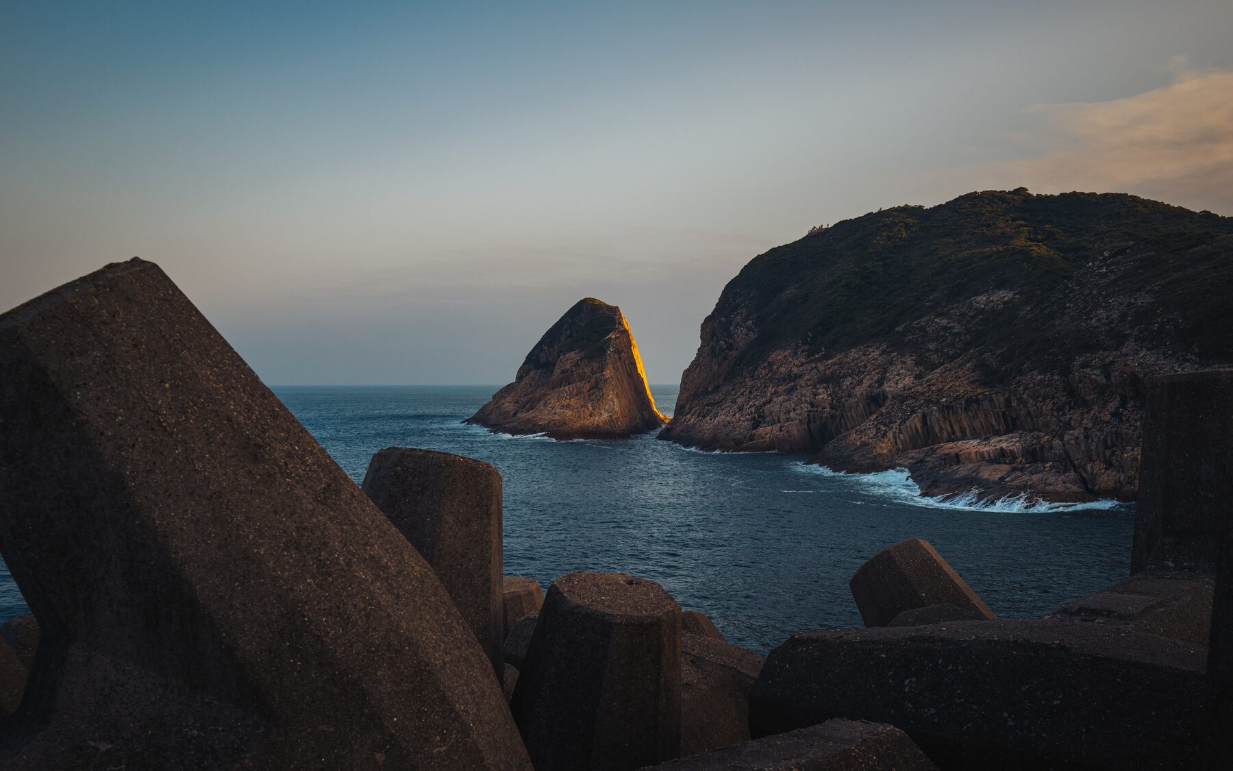A sunlit sea stack and headland at dusk beyond dark concrete blocks.