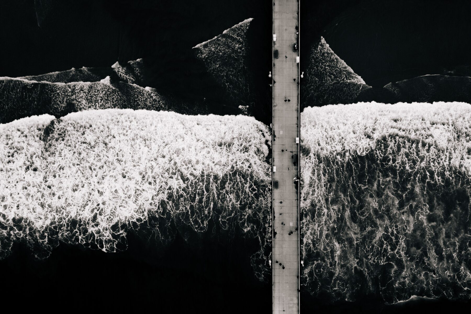 Black and white aerial view of white surf breaking on either side of a narrow pier.